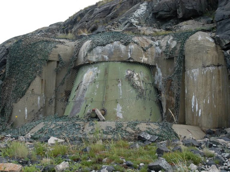 Betongbunker delvis dekket med kamuflasjenett og vegetasjon, bygget inn i en steinete åsside, med en stor, værbitt grønnmalt seksjon foran.