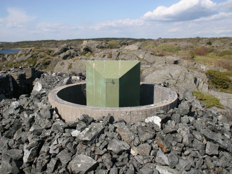 Sirkulær betongstruktur fylt med steiner og grus, med en grønn rektangulær metallgjenstand i midten, omgitt av steinete terreng og utsikt over vann og åser under en delvis skyet himmel.