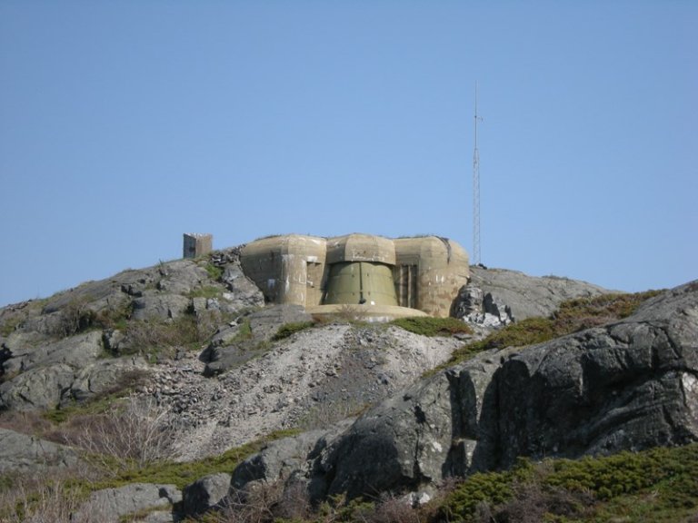 Betongbunker på en steinete høyde med en stor sylinderformet seksjon i midten og mindre seksjoner på sidene, samt en høy antenne til høyre, omgitt av steinete terreng og sparsom vegetasjon.