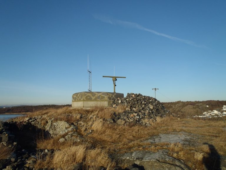 Radarstasjon på en steinete høyde med kamuflasjemønstre, to antenner på toppen og en steinhaug ved siden av, omgitt av tørr gress og steiner under en klar blå himmel.