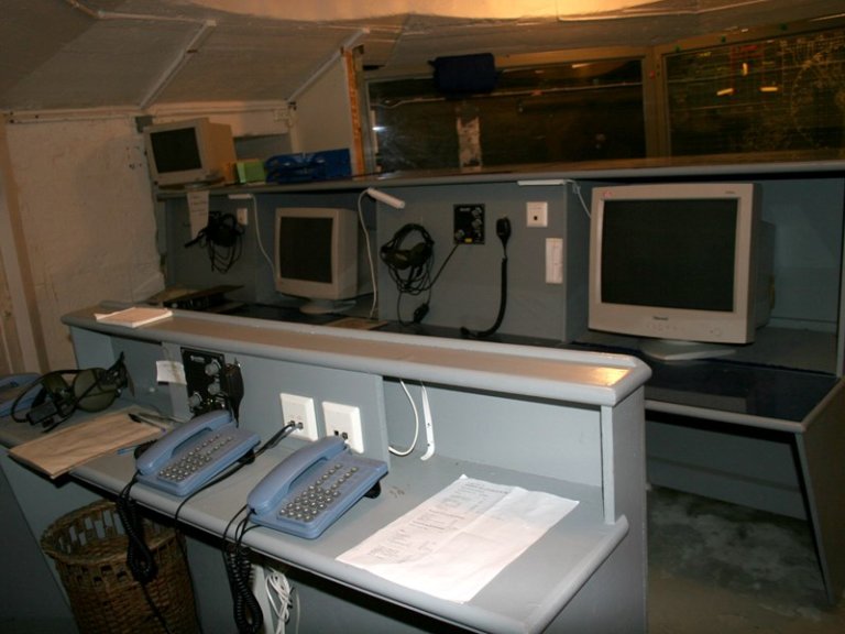 Gammeldags kontrollrom eller kommunikasjonssenter med to pulter, utstyr som telefoner, hodetelefoner og CRT-skjermer, samt papirarbeid og kontorrekvisita, med et stort vindu eller skjerm i bakgrunnen.