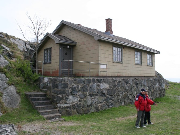 Lite hus bygget på steinfundament med beige kledning, grått tak og pipe, med trapp opp til inngangen og to personer i røde jakker foran huset på gresset.