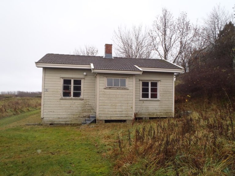 Liten, værbitt trehus med takstein og mursteinspipe, omgitt av gress og overgrodd vegetasjon, med nakne trær i bakgrunnen.