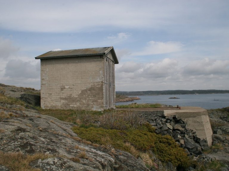 Liten, værbitt betongbygning med skråtak på steinete kystterreng, med utsikt over vann og landmasser i bakgrunnen under en delvis skyet himmel.