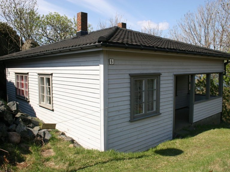 Liten, hvit trehus med svart takstein, to piper, tre vinduer og en åpen veranda, omgitt av gress og steiner, med trær uten blader i bakgrunnen.