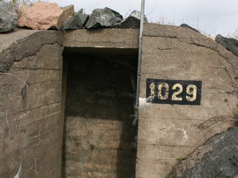 Inngang til betongbunker med værbitt betong, synlige sprekker og steiner på toppen, med et metallrør på høyre side og et svart skilt med hvite tall '1029' ved inngangen.