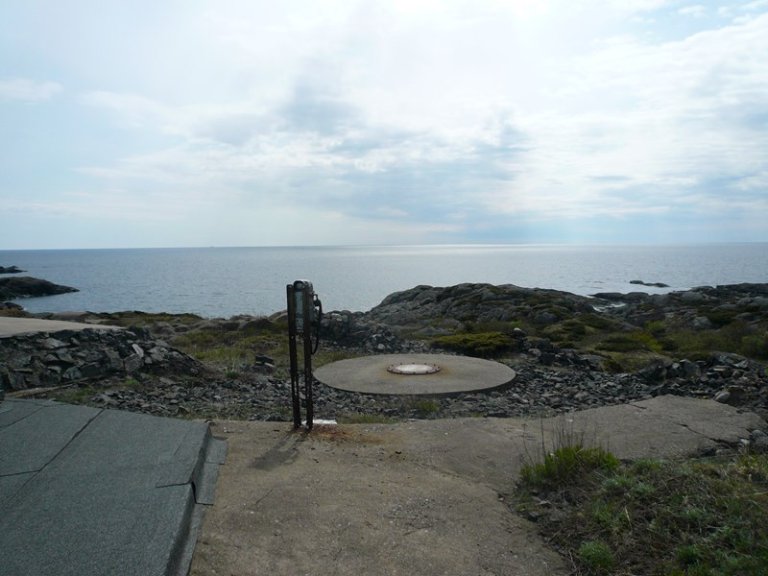 Kystscene med steinete terreng som fører til havet, med en sirkulær betongstruktur og metalldeksel i forgrunnen, og et vertikalt metallstolpe med utstyr ved siden av, under en delvis skyet himmel.