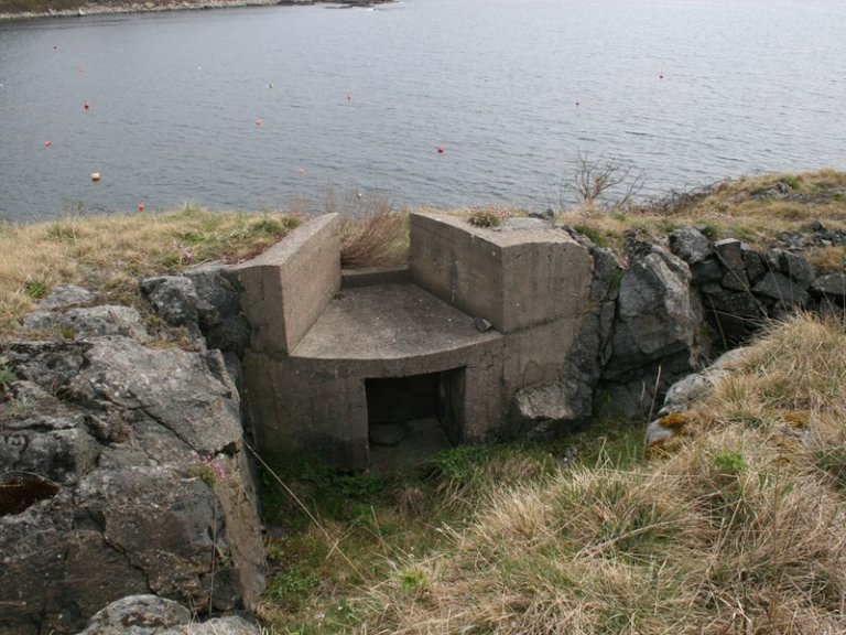 Betongbunker på en gresskledd, steinete åsside med utsikt over vann, omgitt av tørket gress og bøyer i bakgrunnen.
